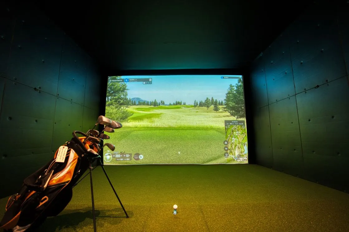 Indoor golf simulator bay with a projected course on screen and a golf bag on turf