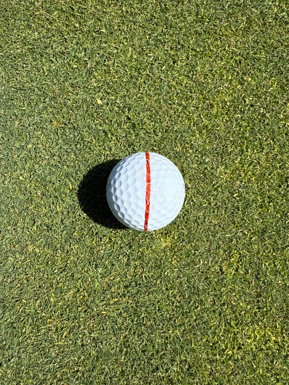 Top-down view of a white golf ball with a red alignment line on a sunlit green