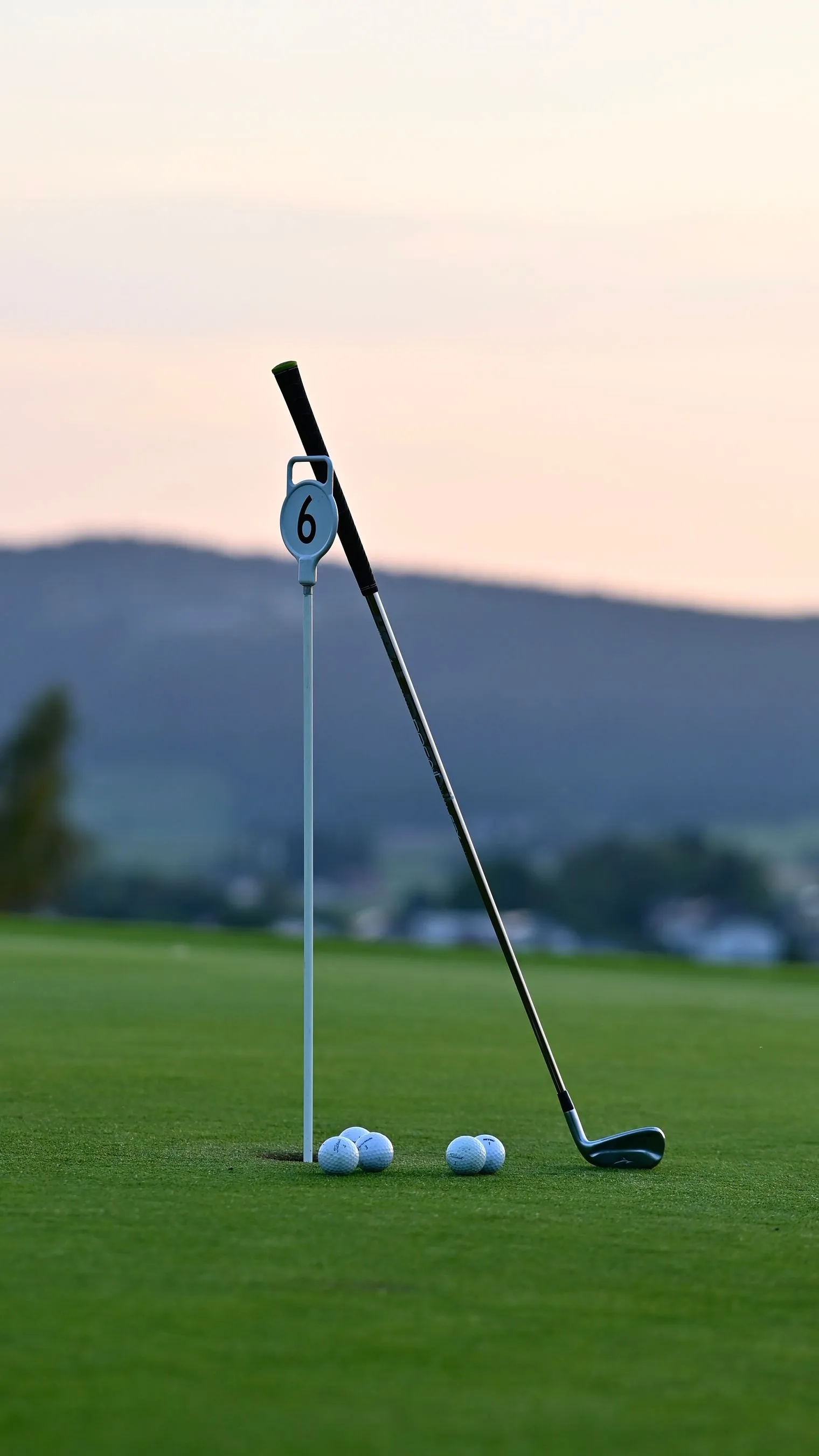 Iron leaning on a flagstick beside golf balls at the cup on a green at sunrise