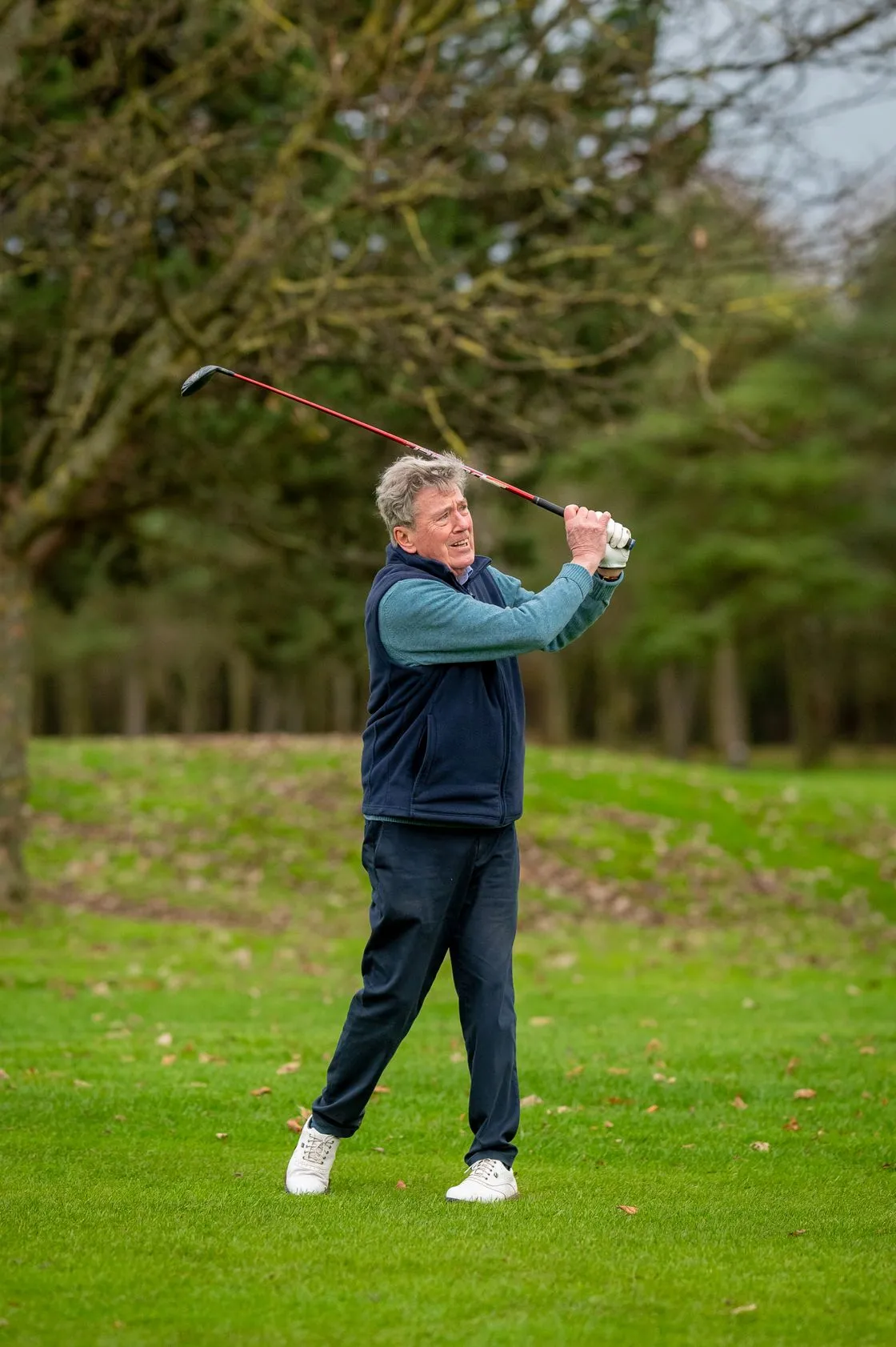 Older golfer in a navy vest finishing a swing on a tree-lined fairway