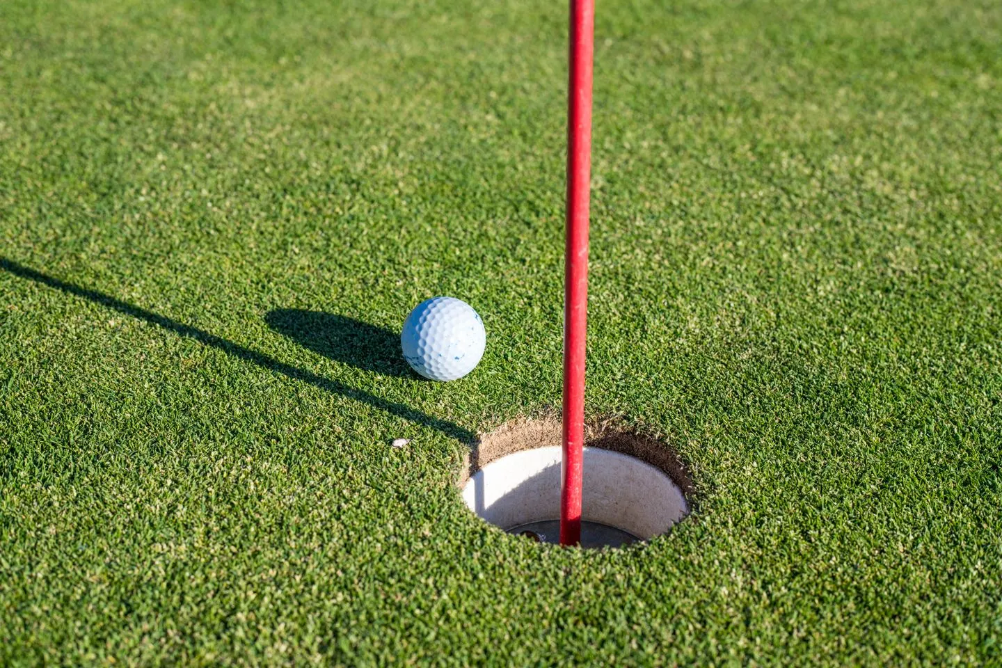 Golf ball resting at the edge of the cup beside a red flagstick on a sunny putting green