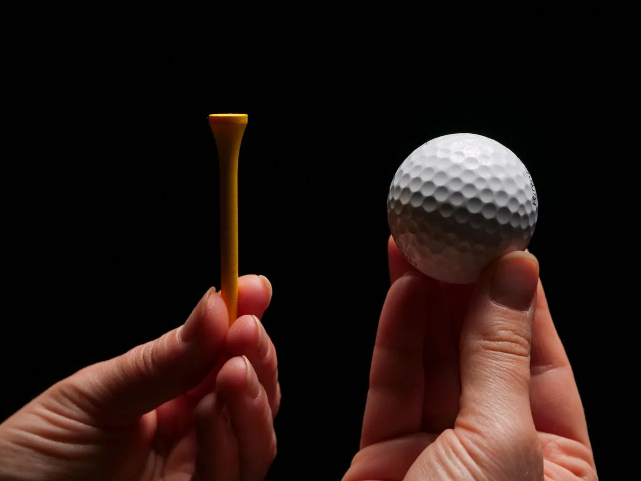Two hands holding up a plain white golf ball and a yellow wooden tee against a black background